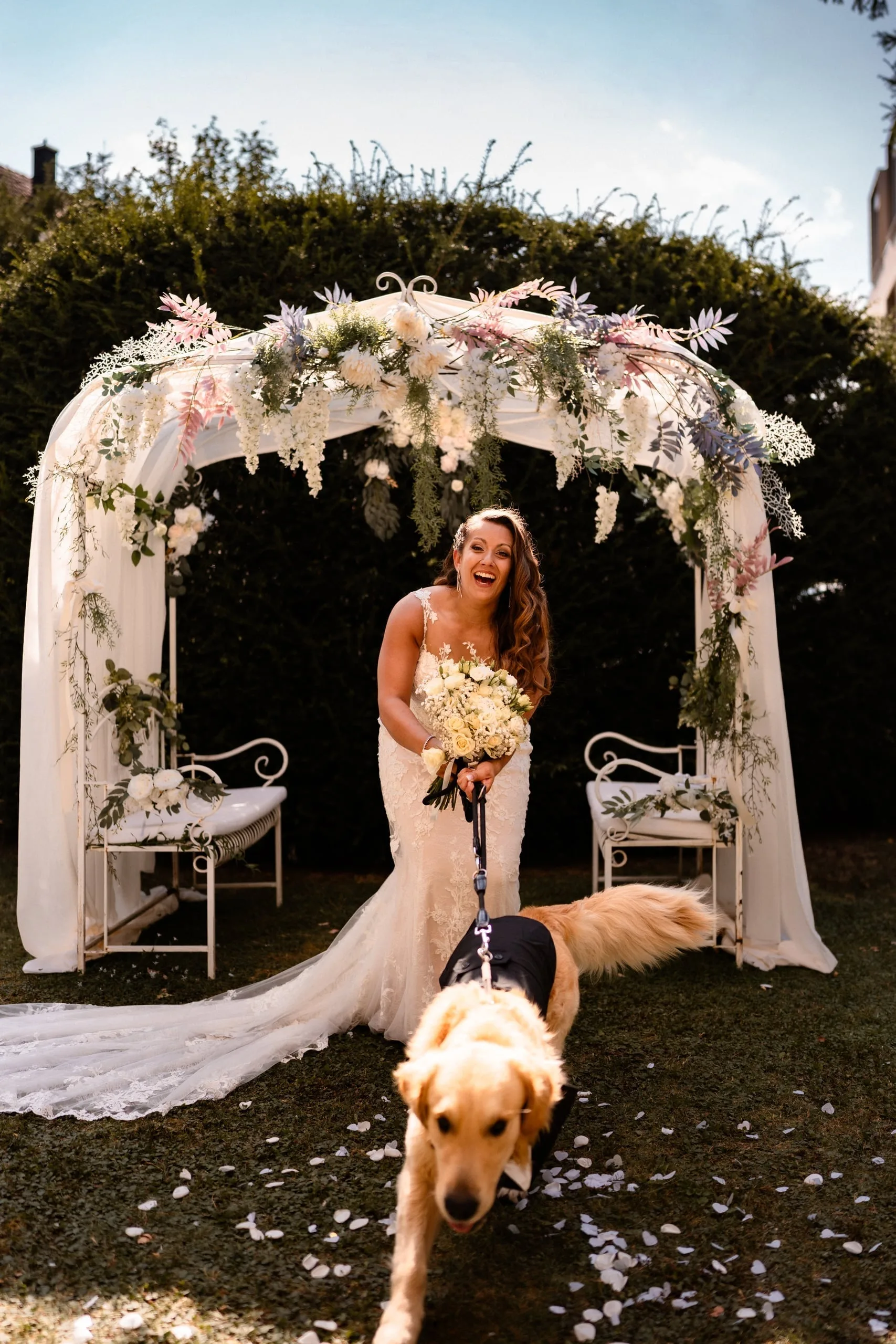 Lachende Braut unter dem Blumenbogen, ihr Hund zieht an der Leine