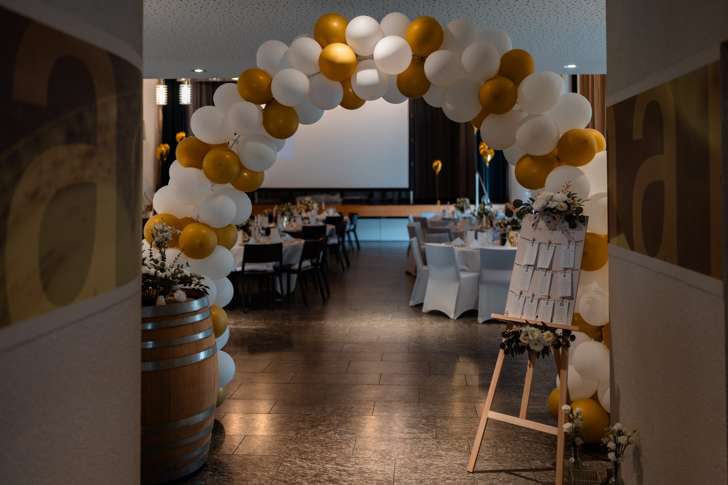 Festlich gedeckter Bankettsaal im B12 mit rund gedeckten Tischen und weiss-goldenem Ballonbogen