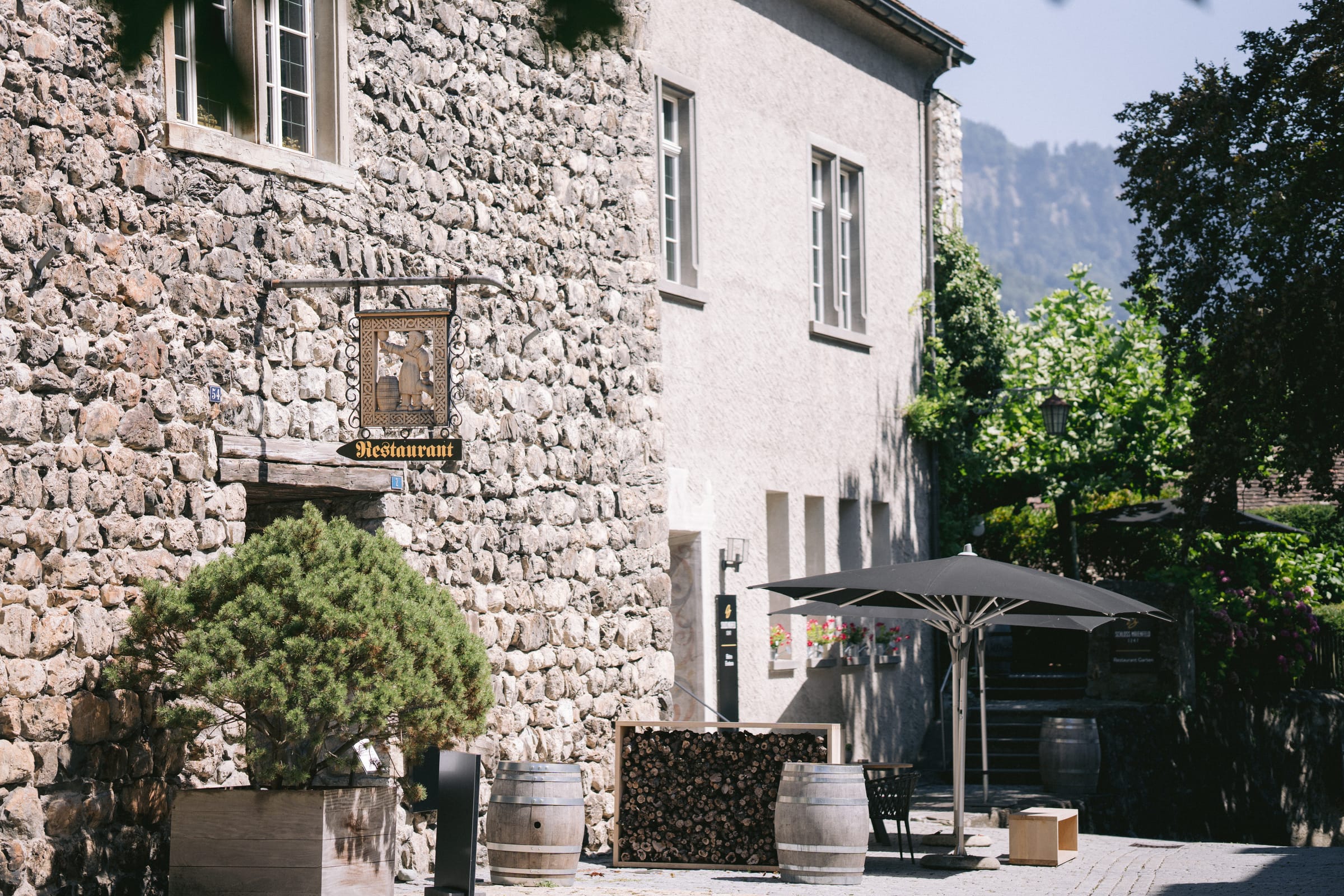 Aussenbereich von Schloss Maienfeld mit schmiedeeisernem Restaurant-Schild, Sonnenschirmen und Weinfässern vor der Bergkulisse