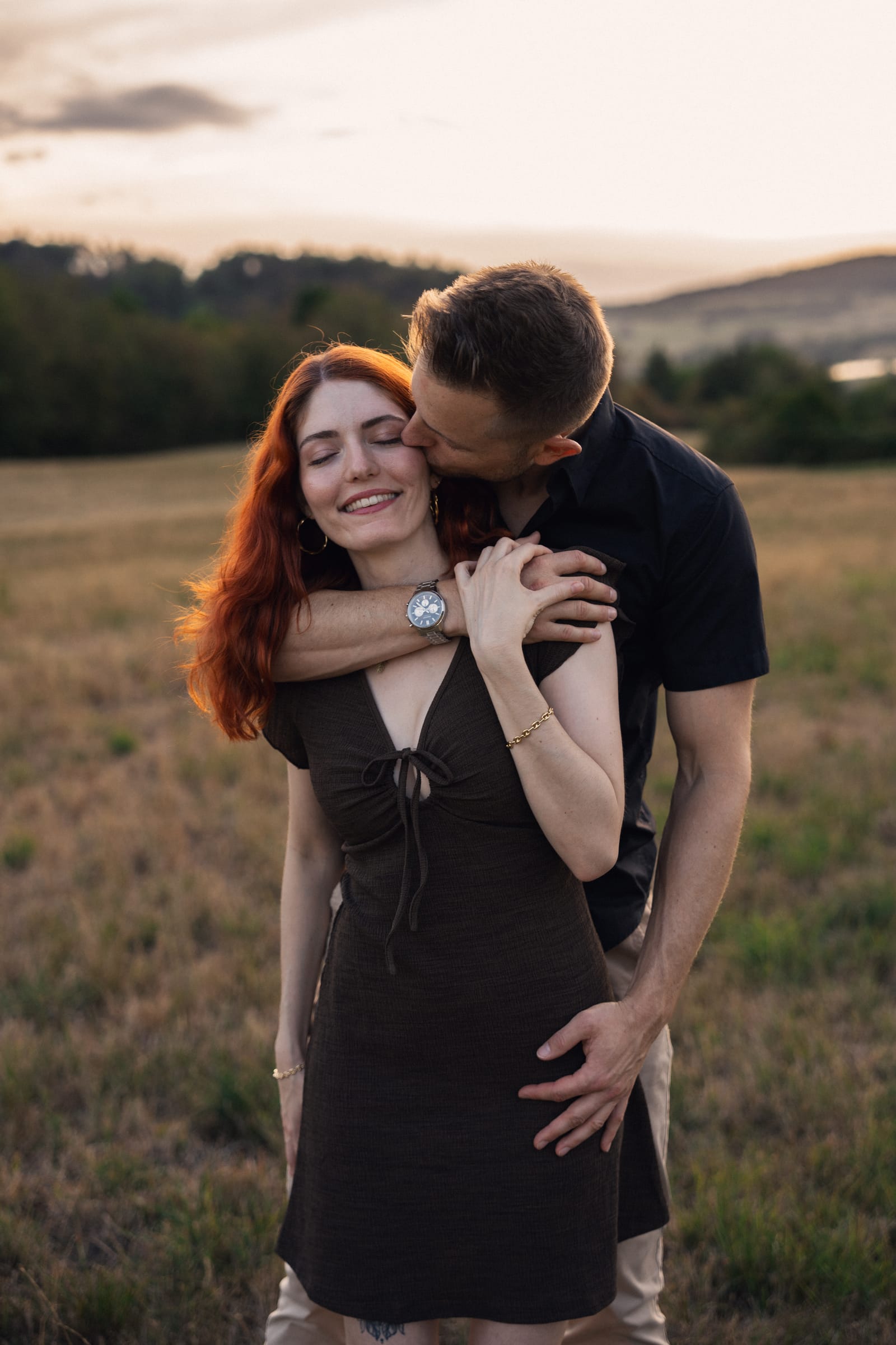 Yanick und Tatjana eng umschlungen auf der trockenen Weide, Hügel und Wald im Hintergrund