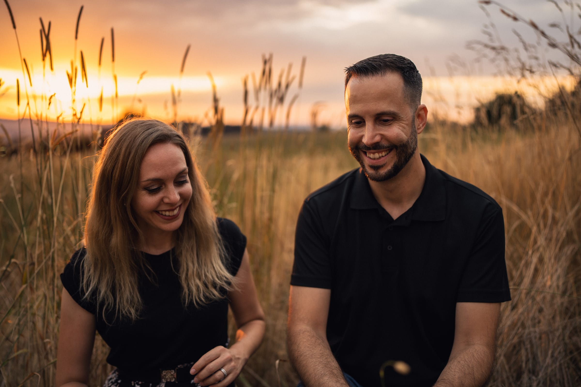 Martina und Stephan sitzen lachend im hohen Gras, die Sonne geht hinter ihnen unter